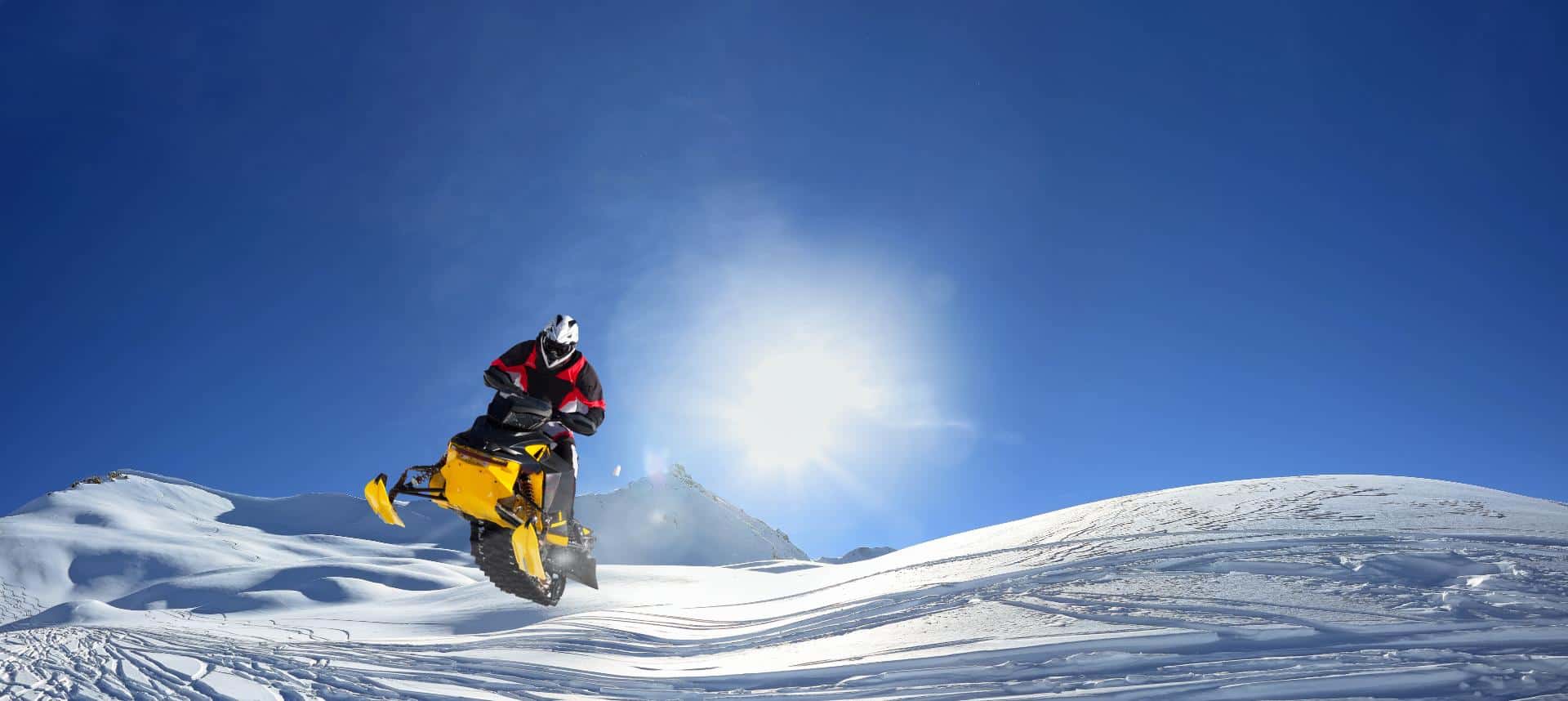 Snowmobile rider climbing a snowy hillside under a bright blue sky with sun overhead