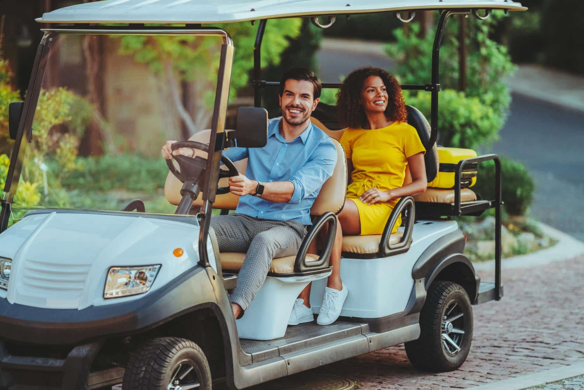 Two people riding in a golf cart along a paved path in a landscaped residential area
